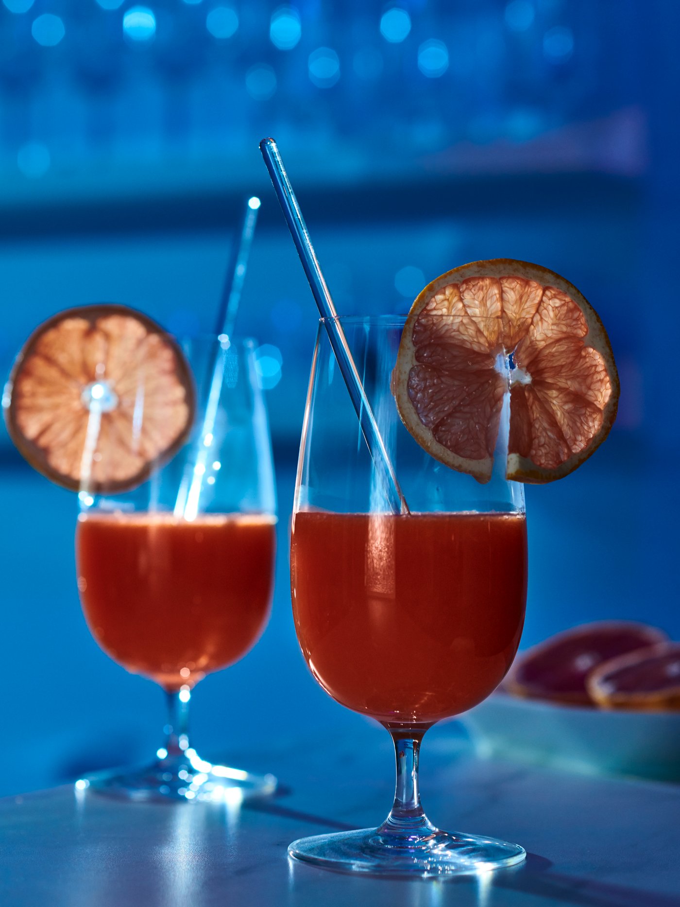 A room bathing in blue light. Two STORSINT beer glasses with stems with blood orange juice and a fruit slice on each rim.