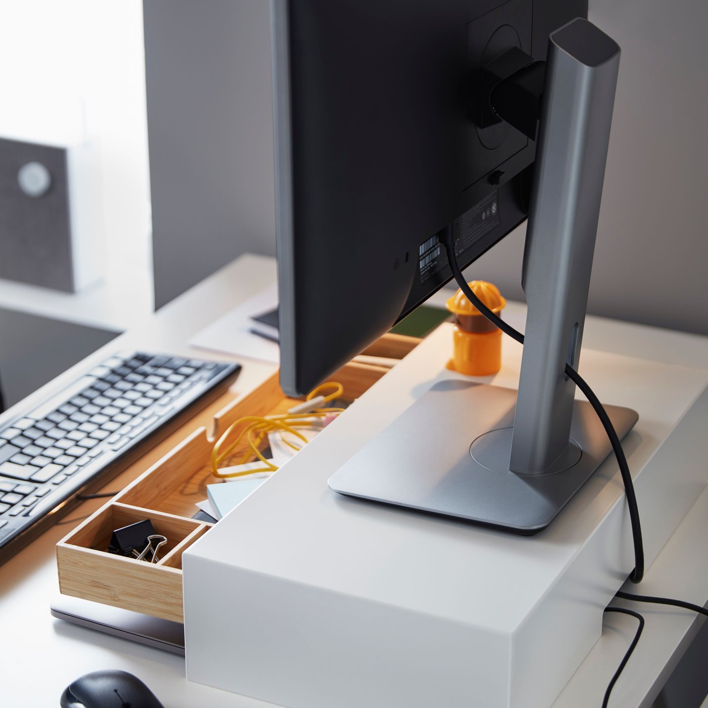 A white ELLOVEN monitor stand with drawer stands on a desk, with a monitor on top of it. The drawer is open.