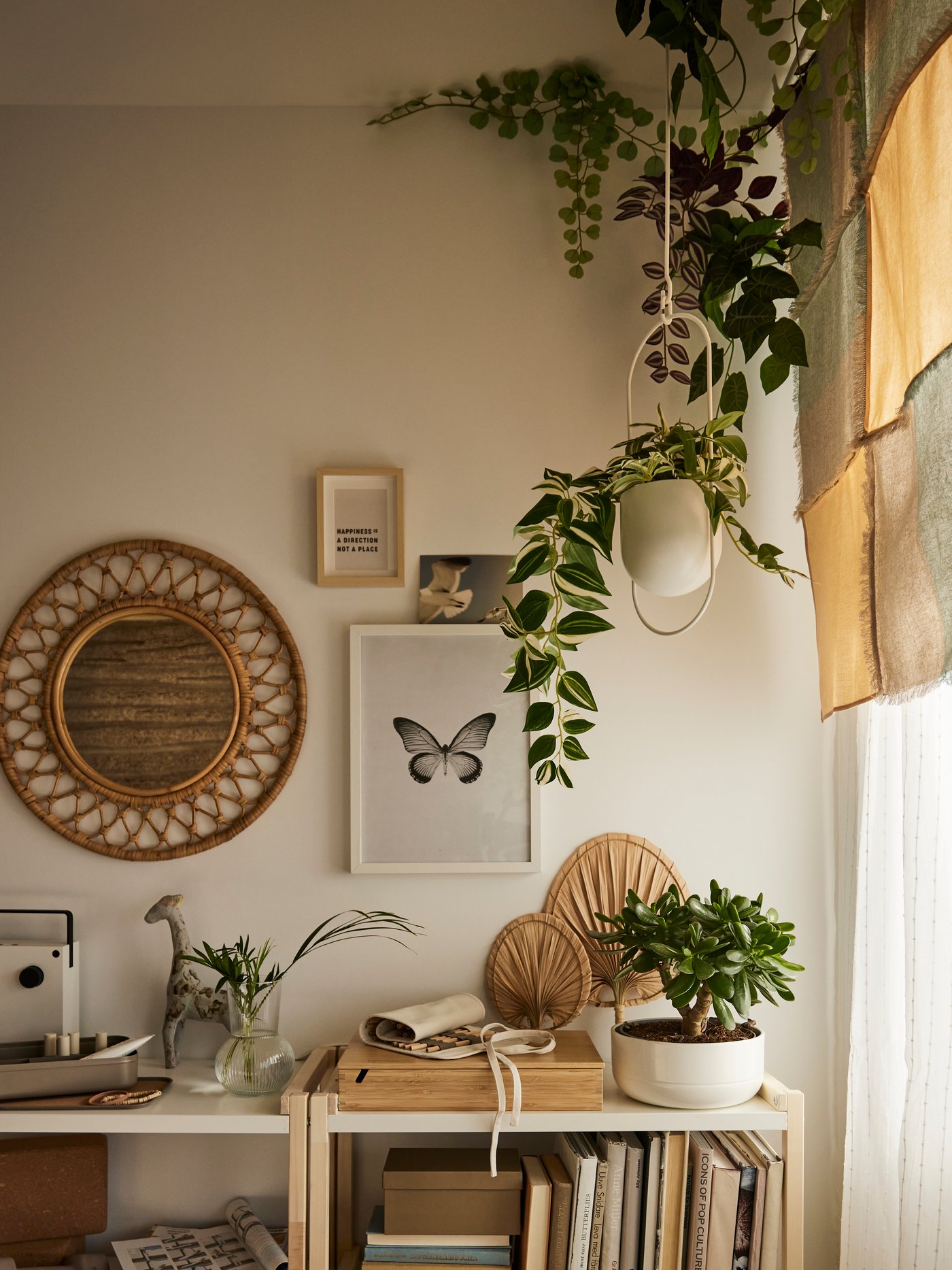 Es ist die Ecke eines Schlafzimmers im Boho-Stil mit einem Rattanspiegel an der Wand, offenen EKENABBEN Regalelementen und Hängepflanzen zu sehen.
