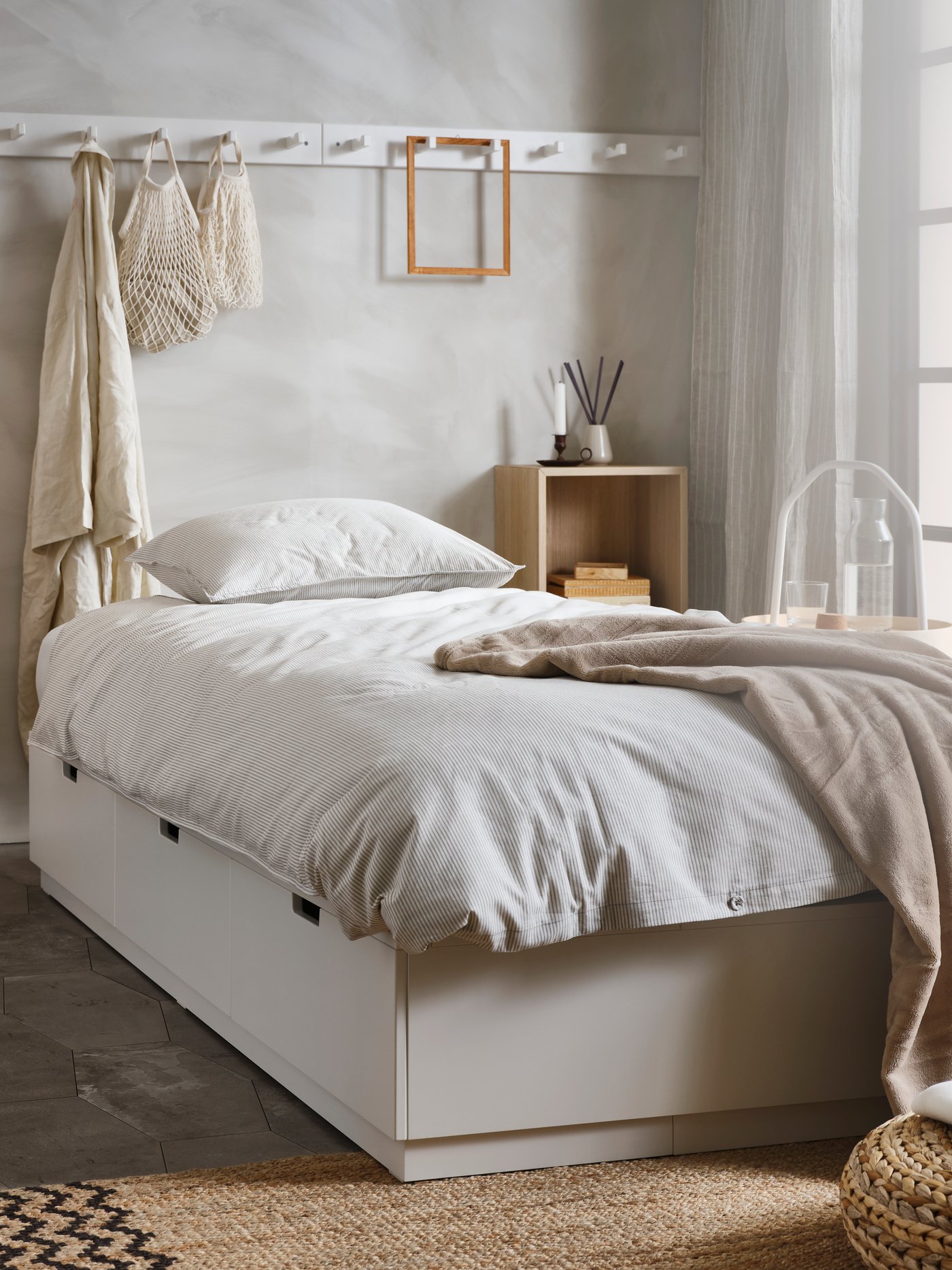 A white NORDLI bed with storage has a wall-mounted EKET shelving unit one side and hooks on the wall behind it.