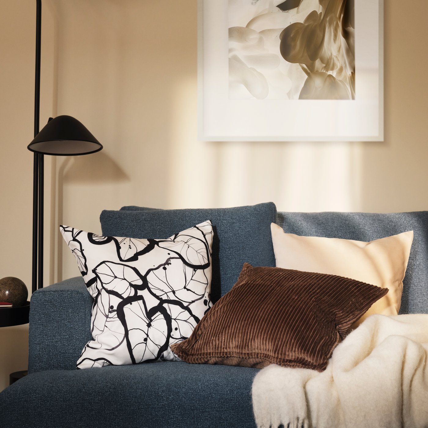 Ein blaues Sofa mit Kissen in verschiedenen Farben und Mustern steht in einem gemütlichen Wohnzimmer. Eine schwarze Stehlampe und ein Wandbild ergänzen die ruhige, stilvolle Einrichtung.