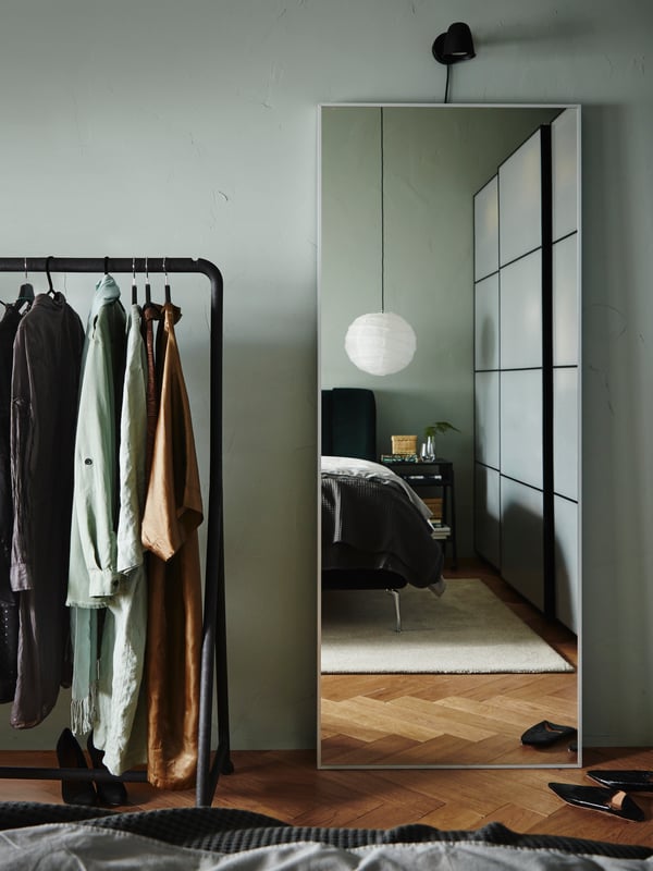 A HOVET mirror against a wall with a SKURUP lamp on the wall above, beside a TURBO clothes rail with hanging garments.