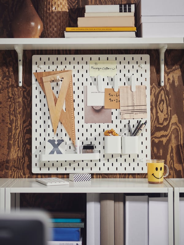 Desk accessories and notes attached to a white SKÅDIS pegboard beneath a white BERGSHULT shelf on a brown wood-panel wall.