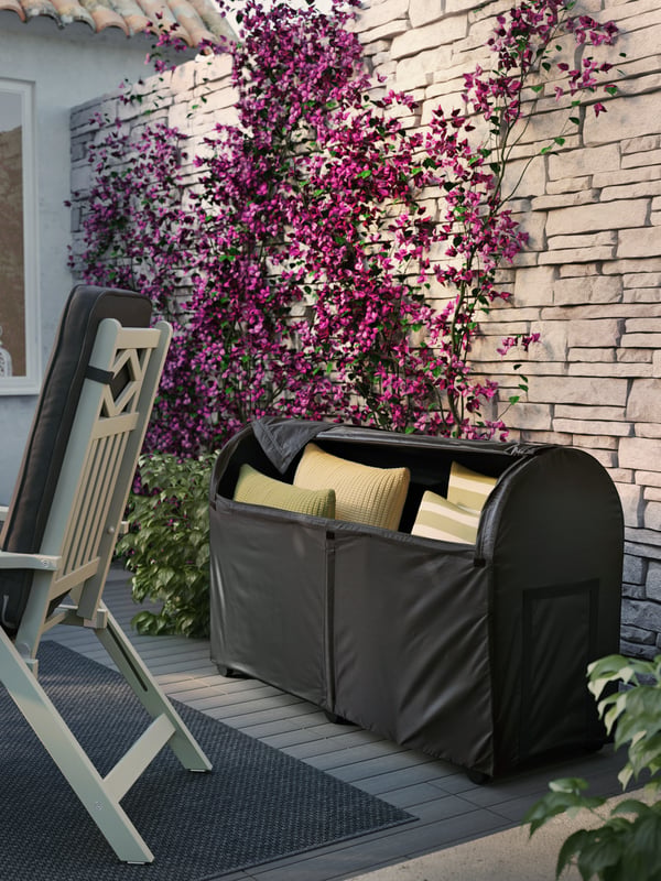 Custodia nera contenente cuscini verdi, appoggiata a un muro di mattoni con rampicante dai fiori viola, dietro a una sedia relax da giardino.