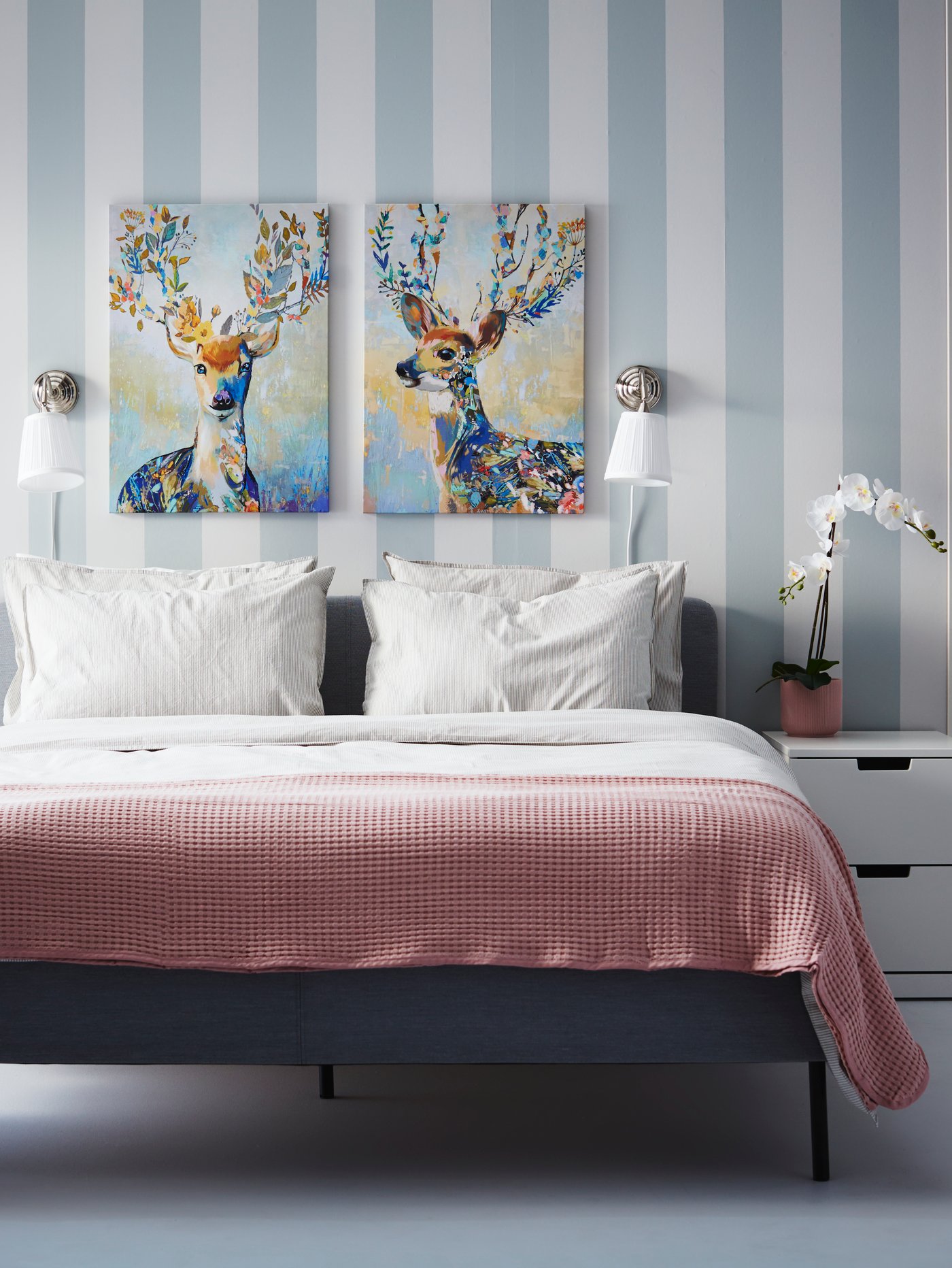 Una cama doble con un plumón rojo con cuadros blancos. En la cabecera de la cama dos cuadros decoraticos con alces dibujados. El papel mural de la habitación es blanco con líneas celestes.
