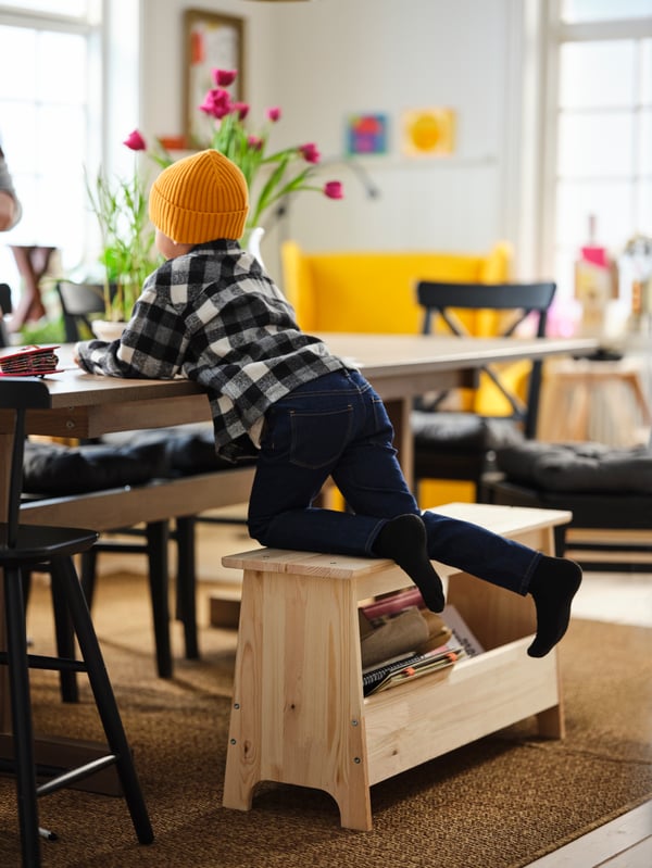 A young child stands on his knees on a PERJOHAN pine bench with storage, his elbows on a grey-brown KLIMPFJÄLL dining table.