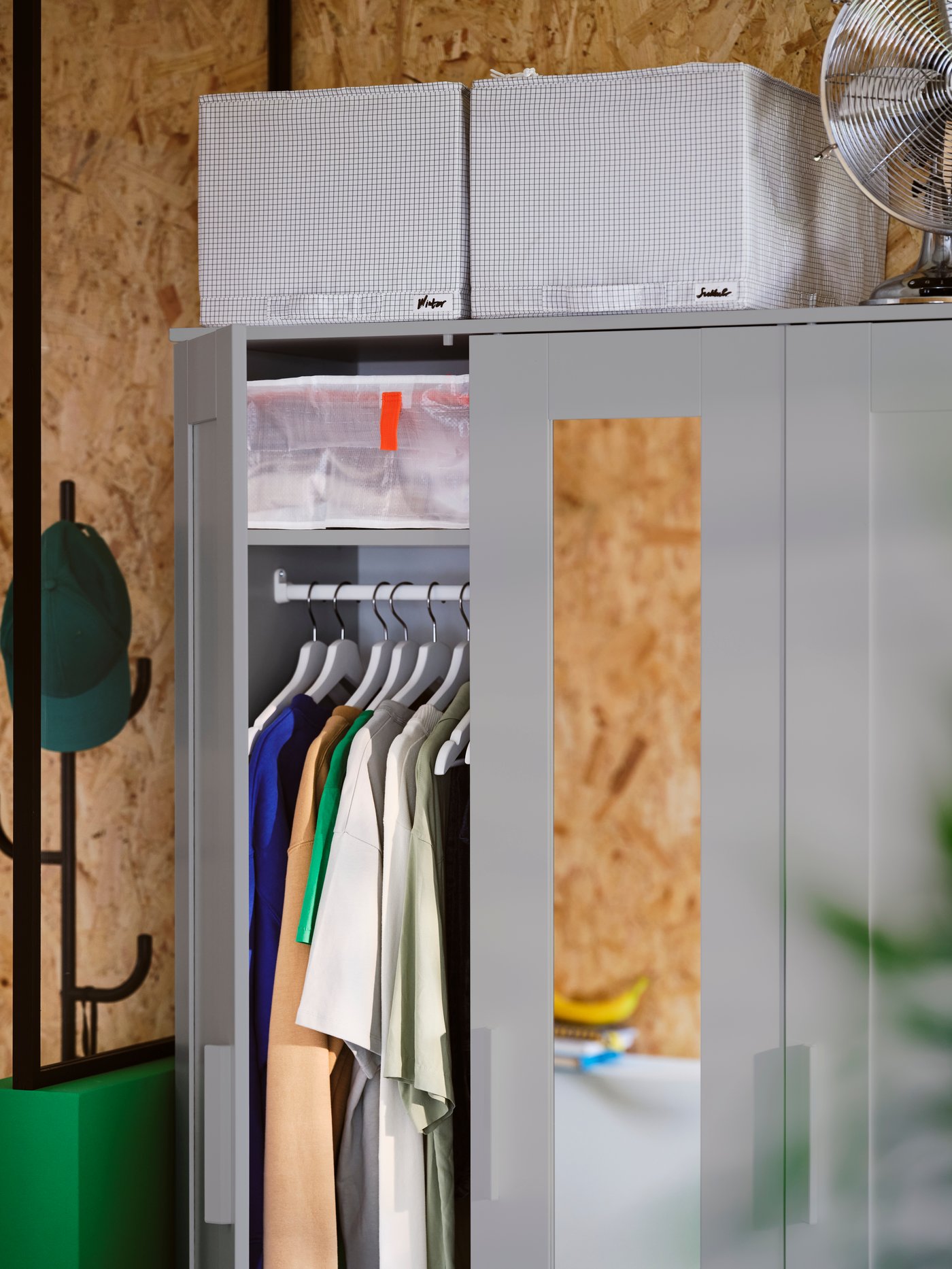 A grey BRIMNES wardrobe with 3 doors has one of its doors open showing hanging clothes. Two STUK storage cases are on top.