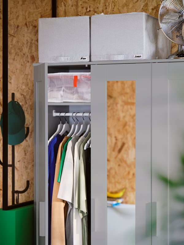 A grey BRIMNES wardrobe with 3 doors has one of its doors open showing hanging clothes. Two STUK storage cases are on top.