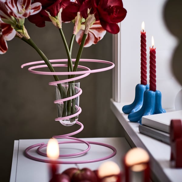 A light pink VINTERFINT vase with a twisted design, containing a transparent vase with amaryllis flowers. On the windowsill, there are two blue VINTERFINT candlestick holders.