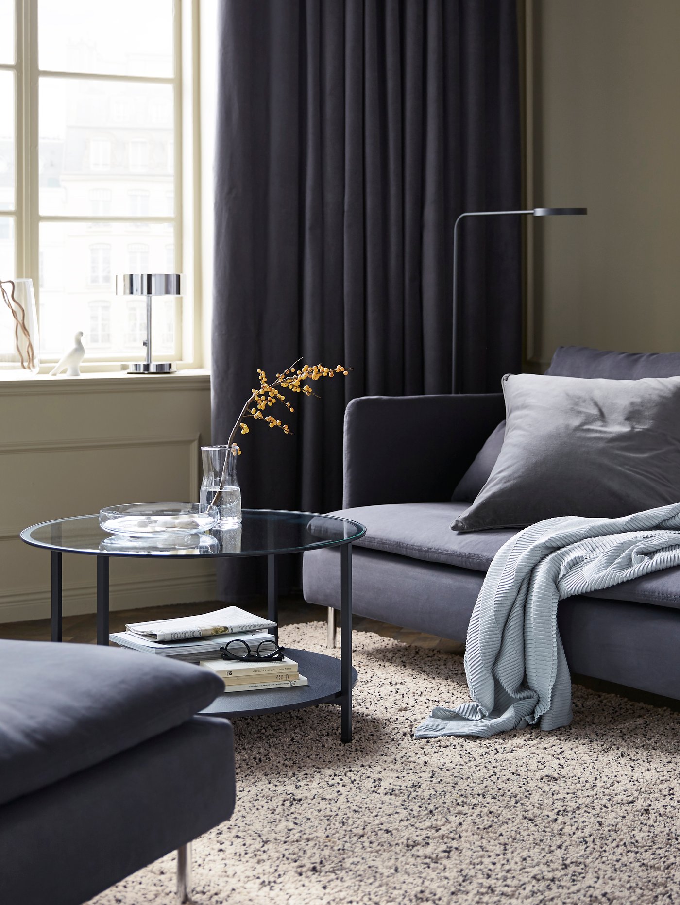 Ein Wohnzimmer mit einem grauen Sofa und grauen Kissen, einem grau-weiß-schwarz gesprenkelten Teppich sowie einem Couchtisch und einer Lampe.