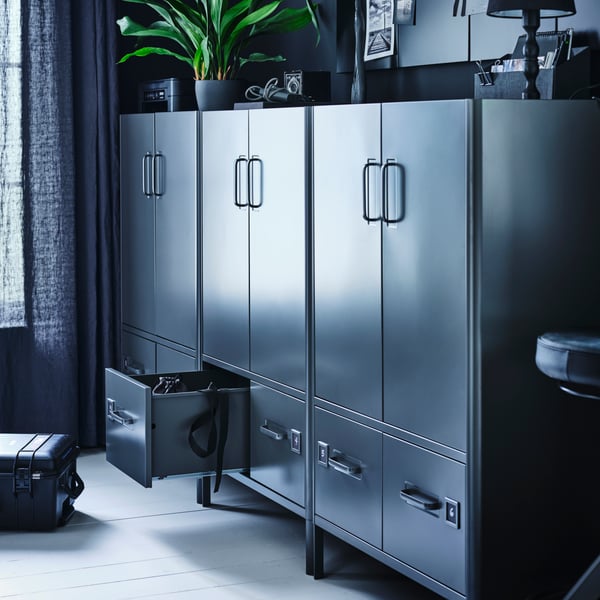 A dark-toned office with three dark grey IDÅSEN cabinets against a wall with a plant on top. One bottom drawer is open.
