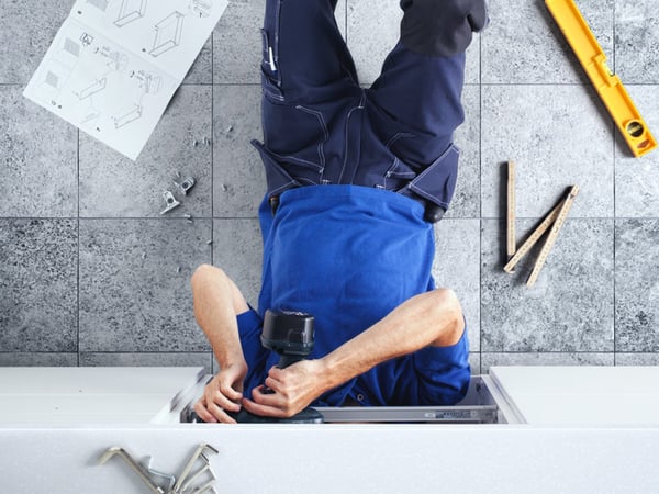 Osoba u plavoj radnoj odeći sastavlja nameštaj, pognute glave, pomoću električne bušilice, a oko nje su po podu s pločicama raštrkani uputstva i alat.