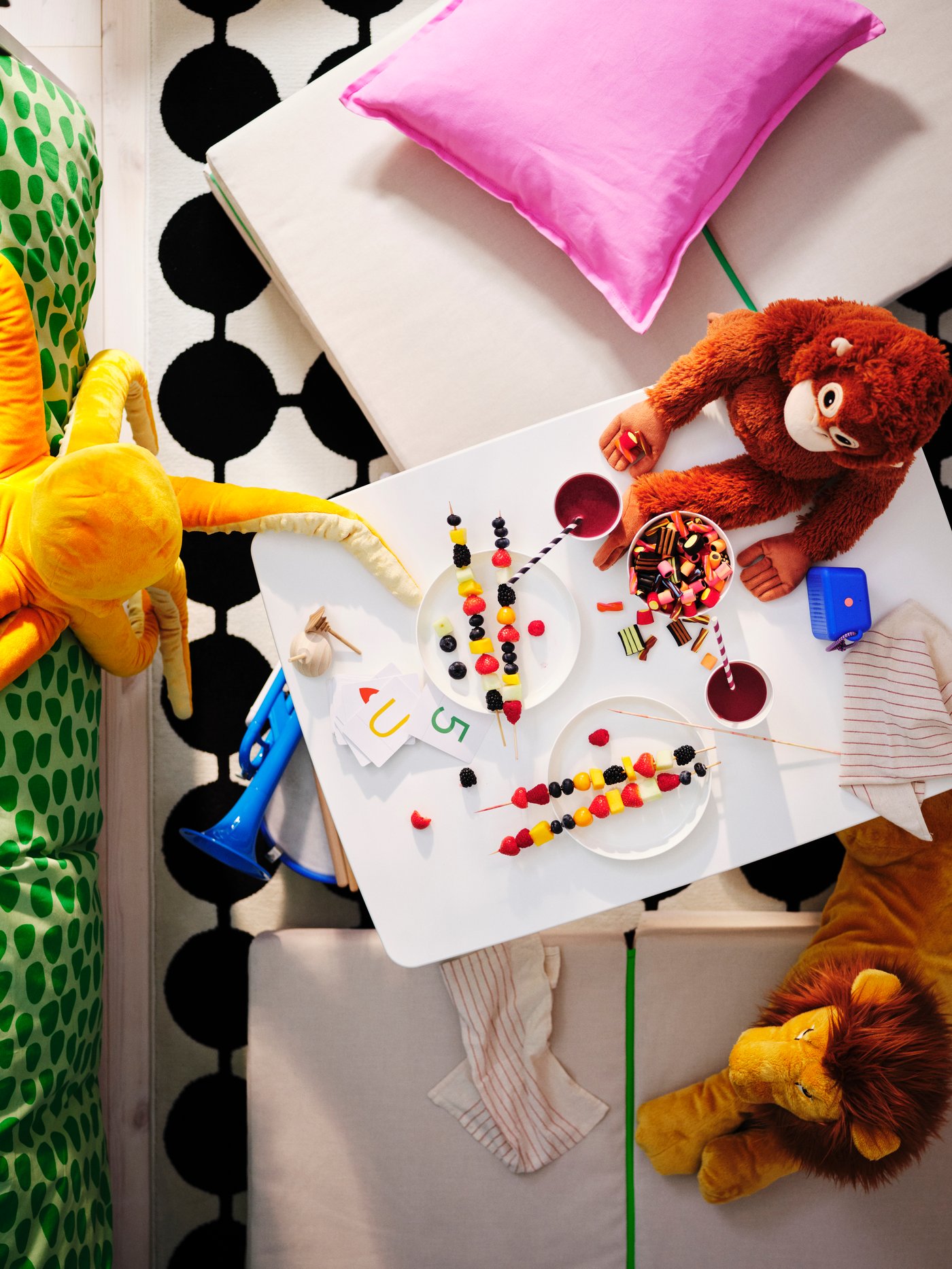 Vue en plongée sur une table MAMMUT sur laquelle sont posés des cartes à jouer, des fruits, des bonbons et des peluches. Un pouf/matelas pliable SLÄKT se trouve au sol, près de la table.