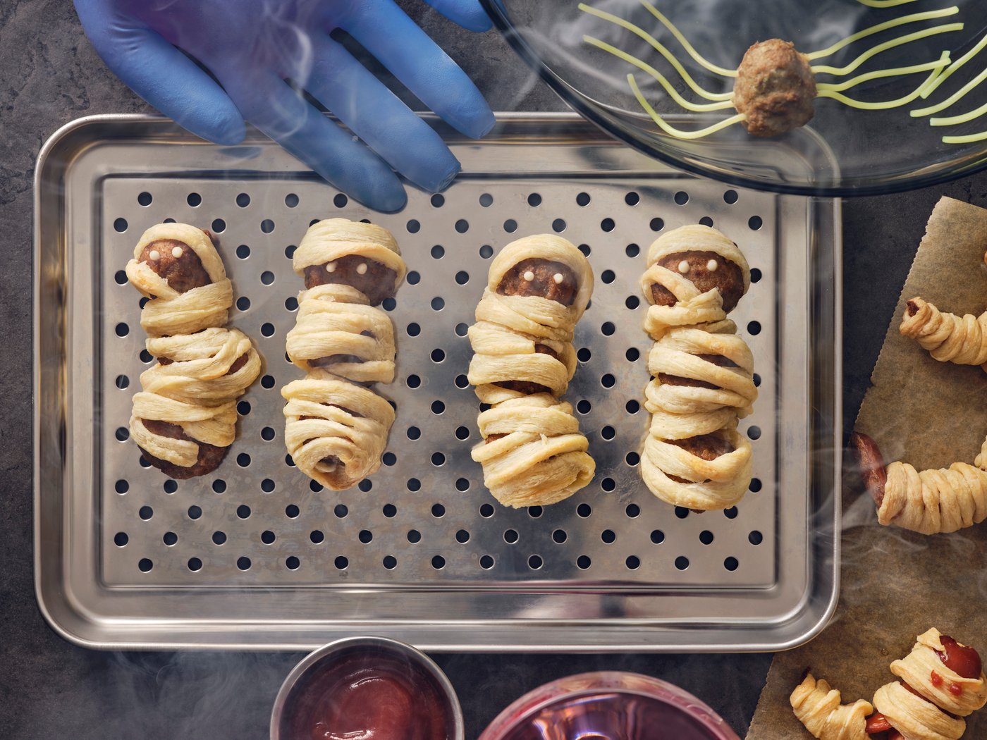 Four meatball mummies made with HUVUDROLL plantballs or meatballs and puffed pastry strips on a stainless steel dish.