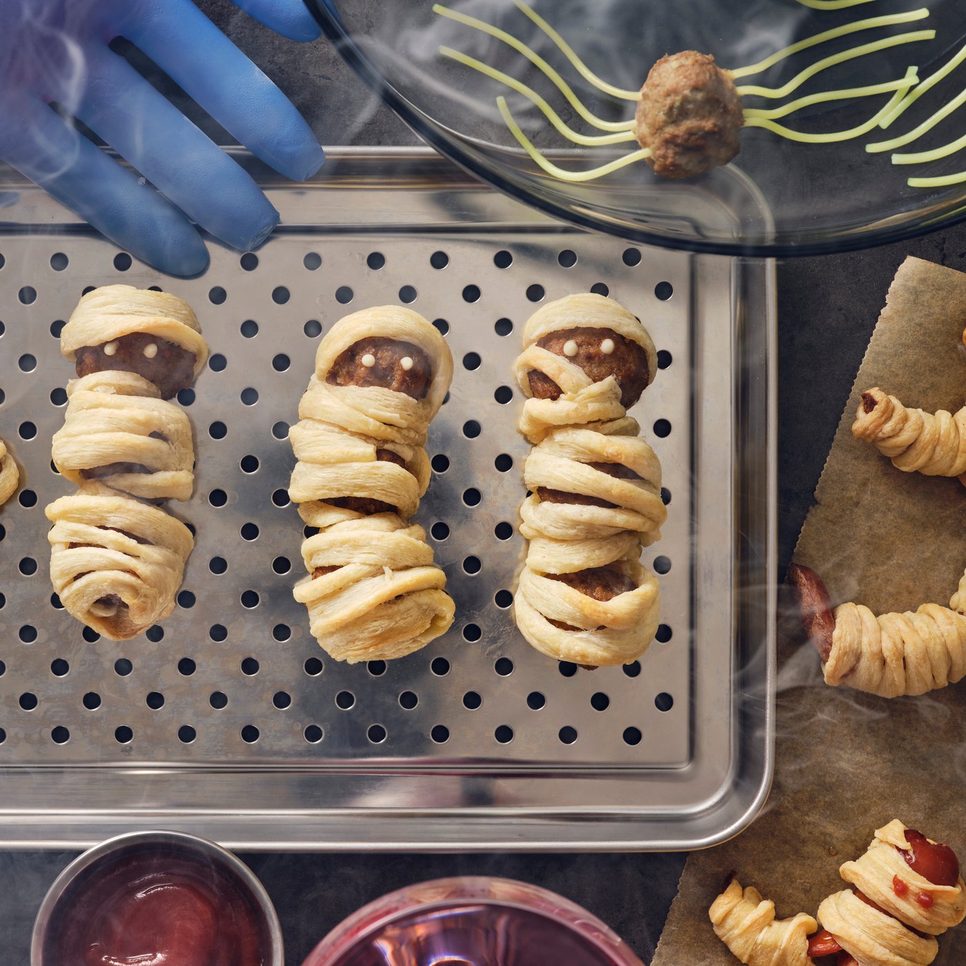 Three meatball mummies made with HUVUDROLL plant balls or meatballs and puffed pastry strips on a stainless steel dish.