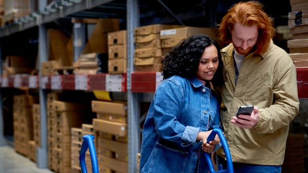 Two people are standing in an IKEA warehouse aisle between tall shelves filled with flat-packed boxes. Both are holding a blue shopping cart and are looking at a smartphone together, suggesting they are using the Shop & Go feature to scan and manage their items.