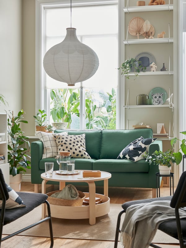 Una habitación iluminada con colores cálidos en la que destaca una mesa de centro BORGEBY y un sillón verde rodeado de plantas.