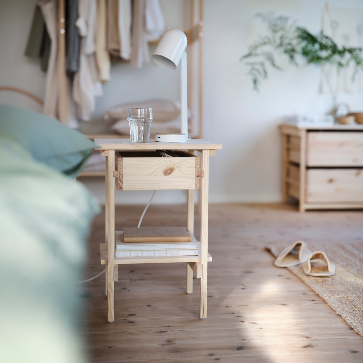 KRONÖREN bedside table in pine with drawer and shelf holding books, placed beside a bed in a light bedroom.