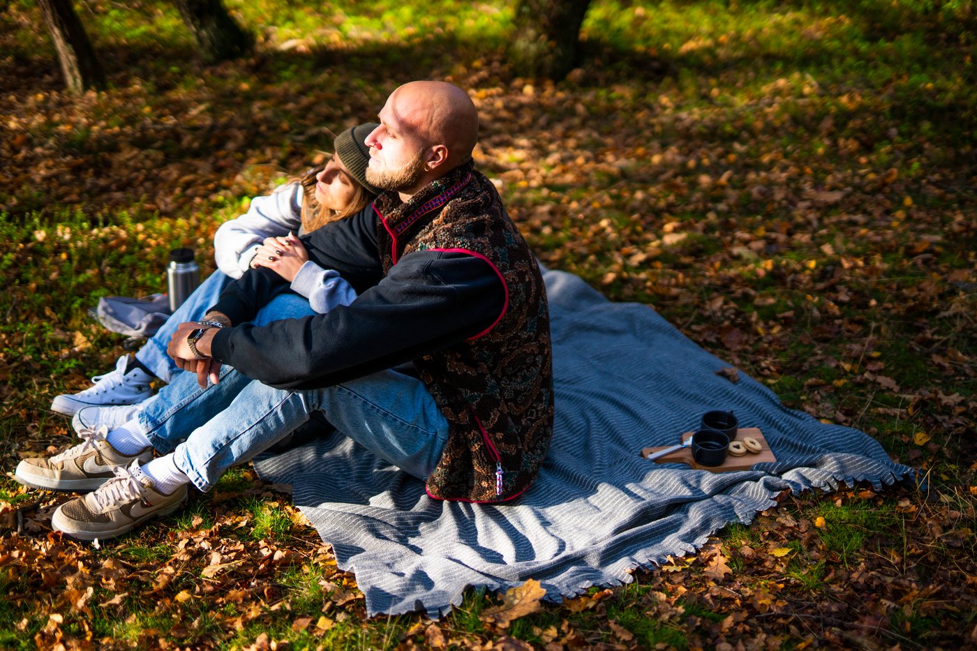 Muškarac i žena sede na izletničkom ćebetu u šumi prekrivenoj suvim jesenjim lišćem.