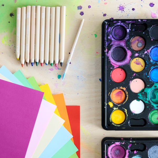Coloured paper, a set of MÅLA pencils and watercolour box, all laying on a wooden table. Picture shot from above.