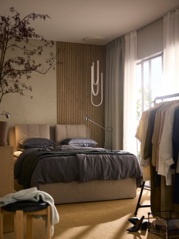 Beige LILLFJÄLLET gestoffeerd bed met opbergruimte, opgemaakt met donkergrijze DYTÅG dekbedovertrekset; erboven hangt een VARMBLIXT ledlamp.