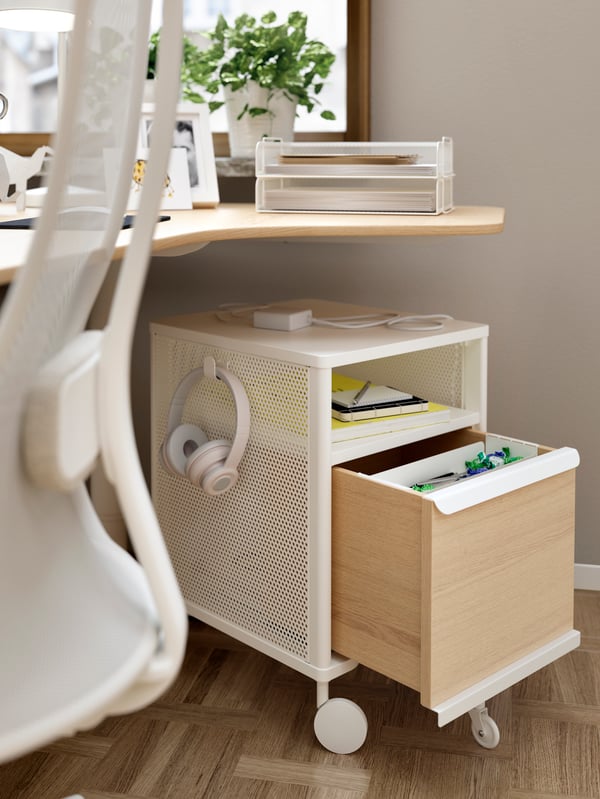 BEKANT storage unit under a desk with an open drawer and objects on the shelf. Headphones hang from a hook on the side.
