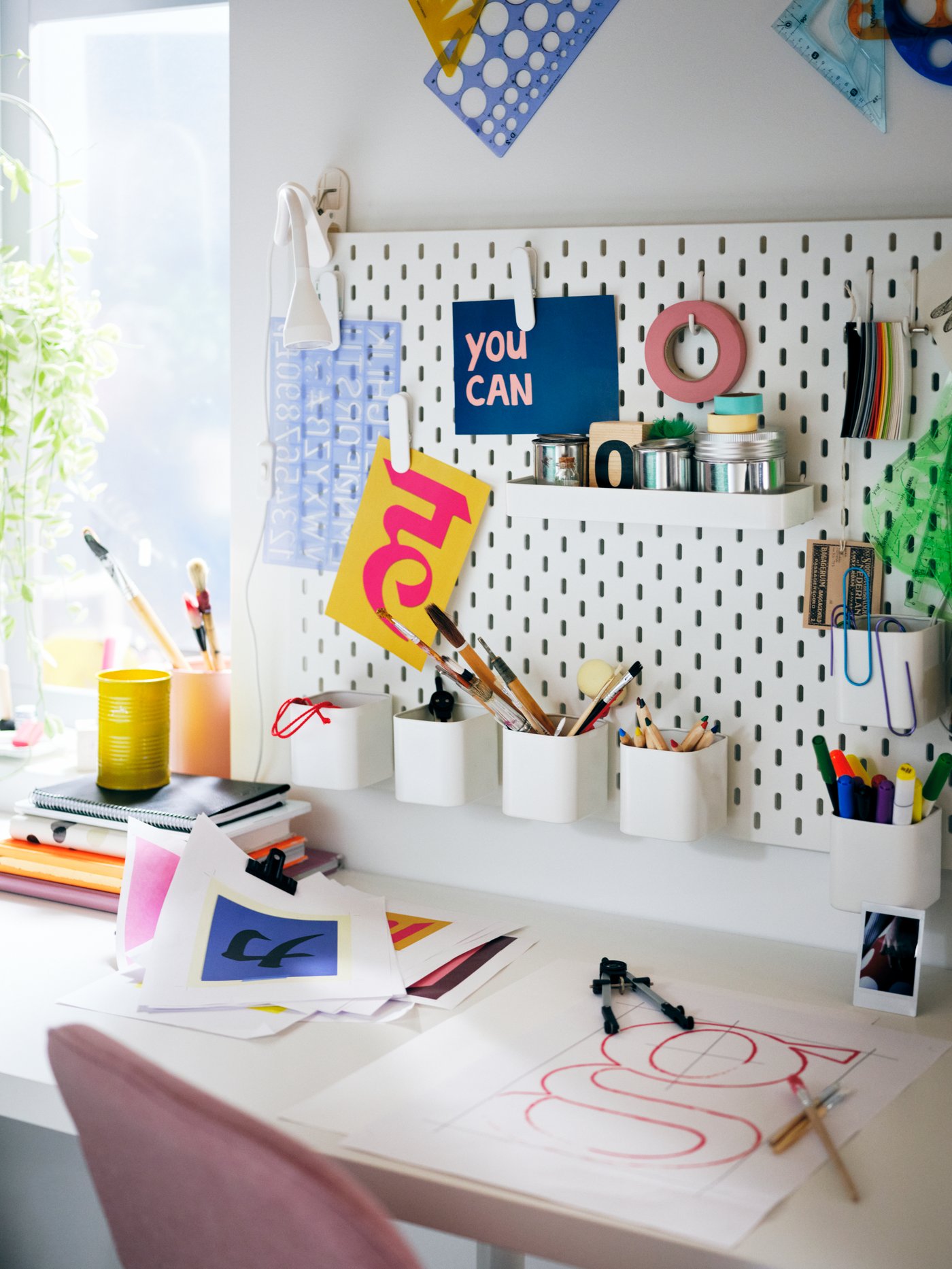 A white SKÅDIS pegboard filled with colourful accessories, over a white LAGKAPTEN tabletop and pink LÅNGFJÄLL swivel chair.