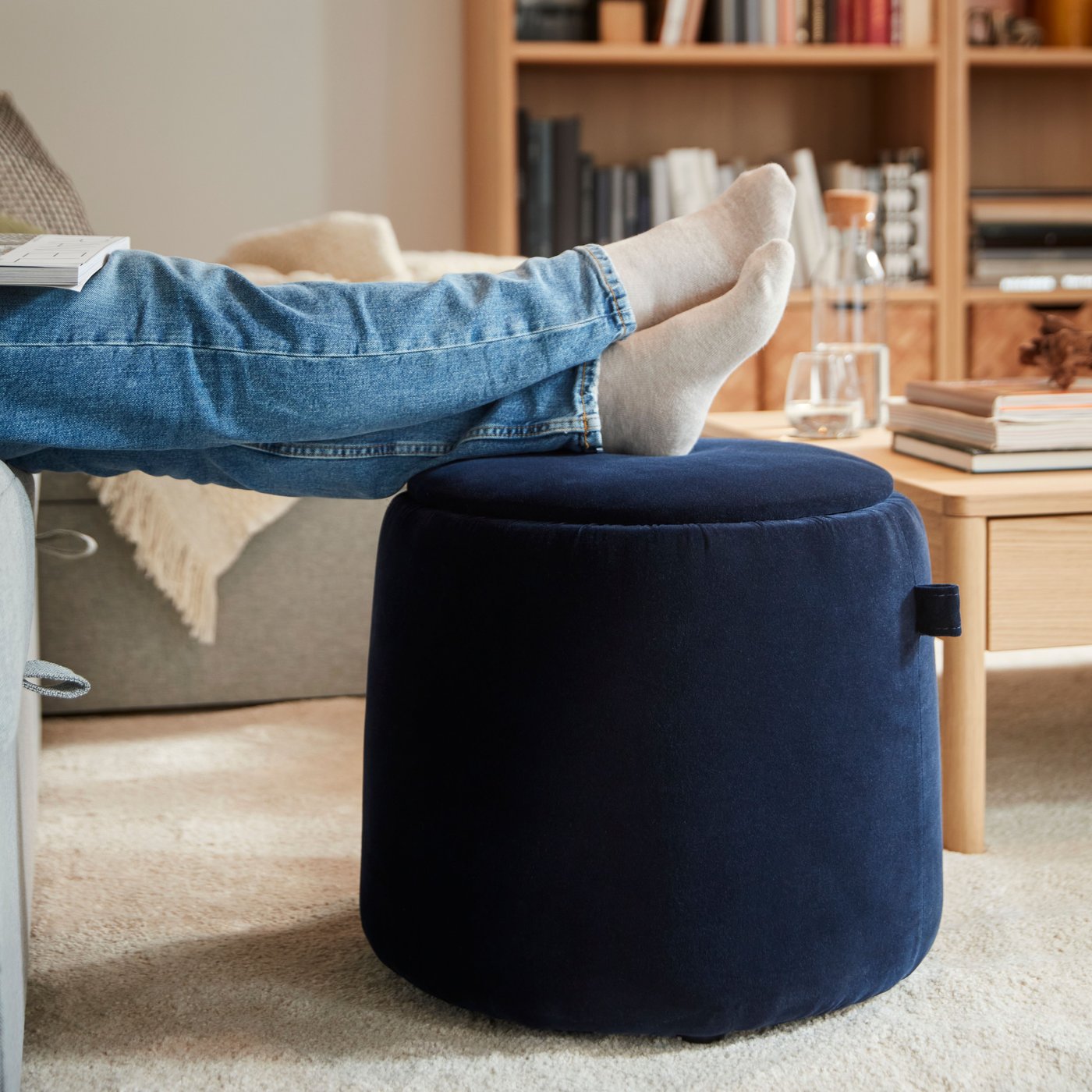 A person’s feet rest on a dark blue JÄRRESTAD pouffe with storage near a TONSTAD bookcase and a TONSTAD coffee table.