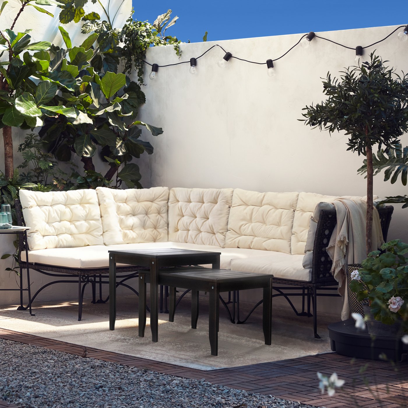 JUTHOLMEN havesofa med beige KUDDARNA hynder står op ad en hvid væg på en terasse fyldt med grønne planter og udendørs lyskæde.