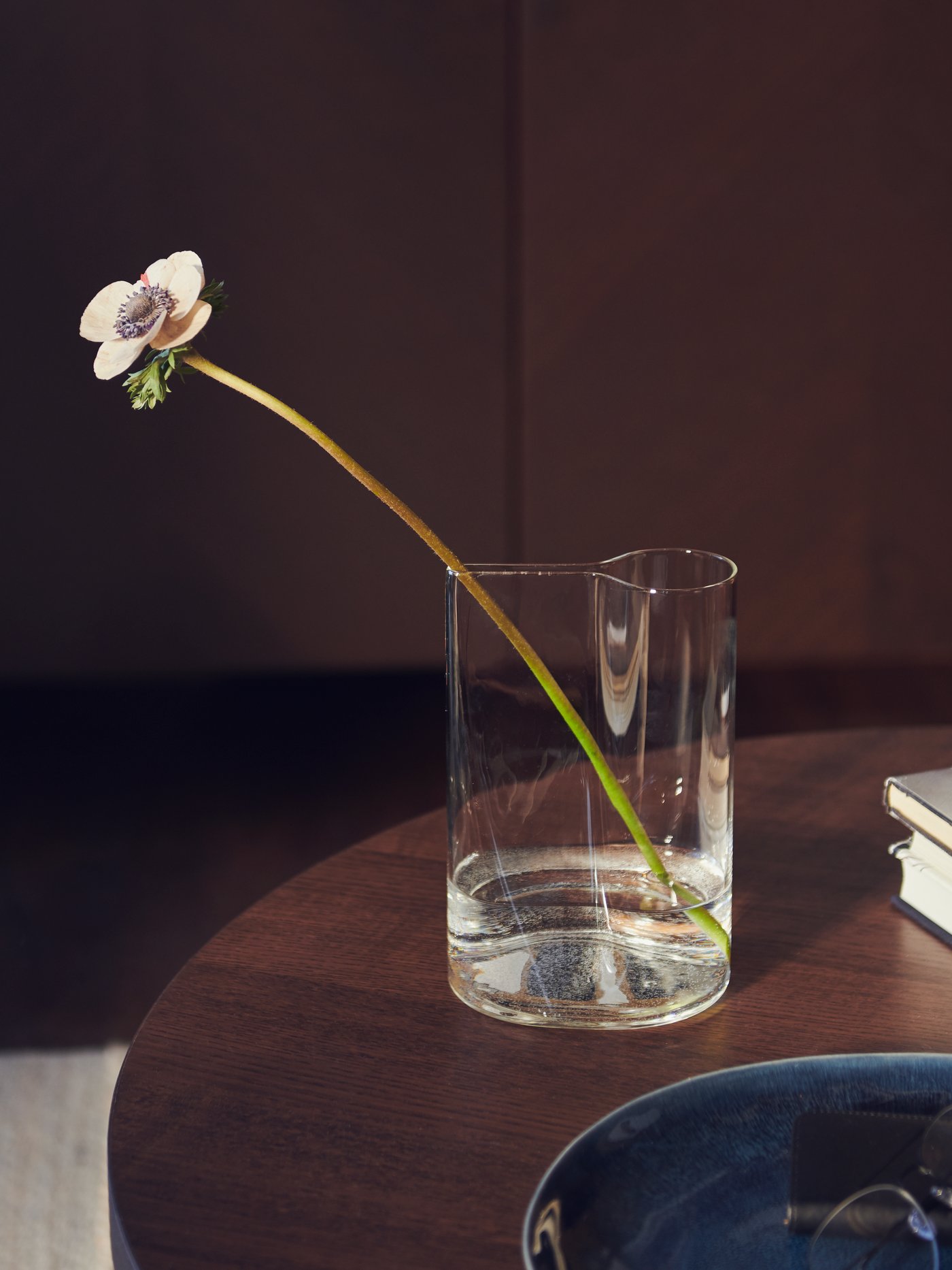 A CHILIFRUKT vase on a curved dark wood table top containing a single flower stem beside a blue bowl and a pile of books.