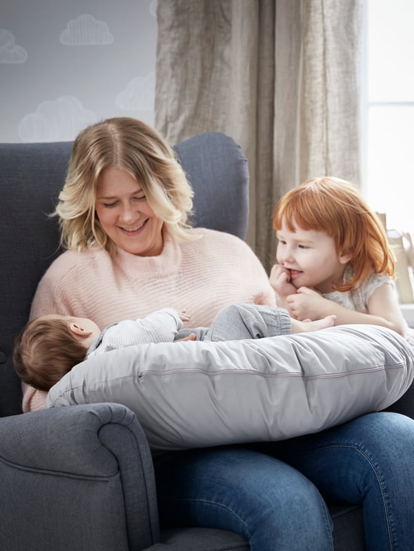 A mother is sitting on a chair holding her baby on a LEN nursing pillow. Beside her is a young child who is smiling.
