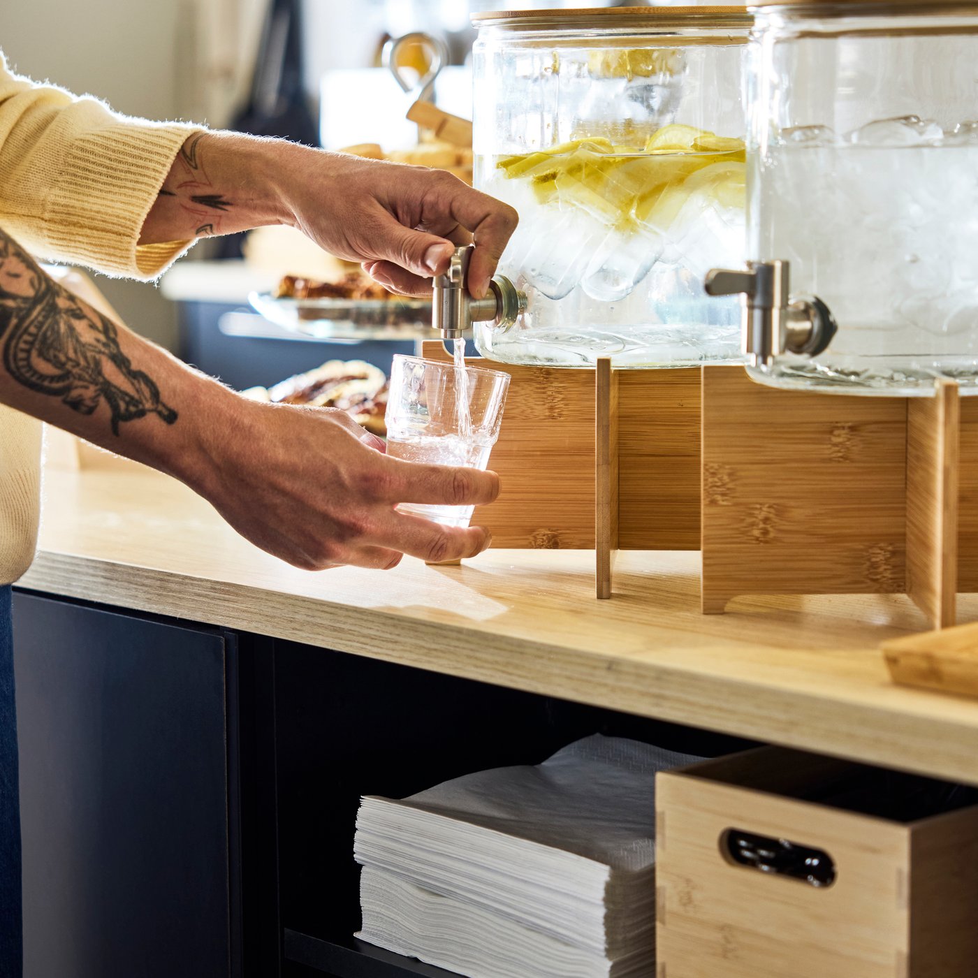 Händerna fyller ett glas från en vattenautomat med citronskivor och is på ett träställ, placerat på en kafédisk.