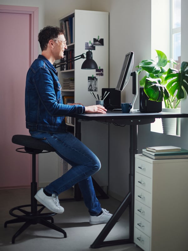 Un homme assis sur un tabouret ergonomique noir devant un bureau réglable regarde l’écran de son ordinateur. 