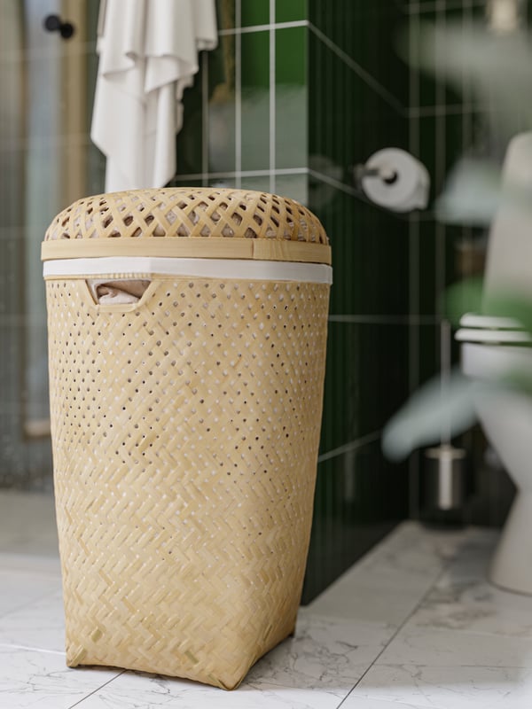 A SALUDING laundry basket of bamboo is standing between a shower and a toilet and there is a white towel hanging above it.