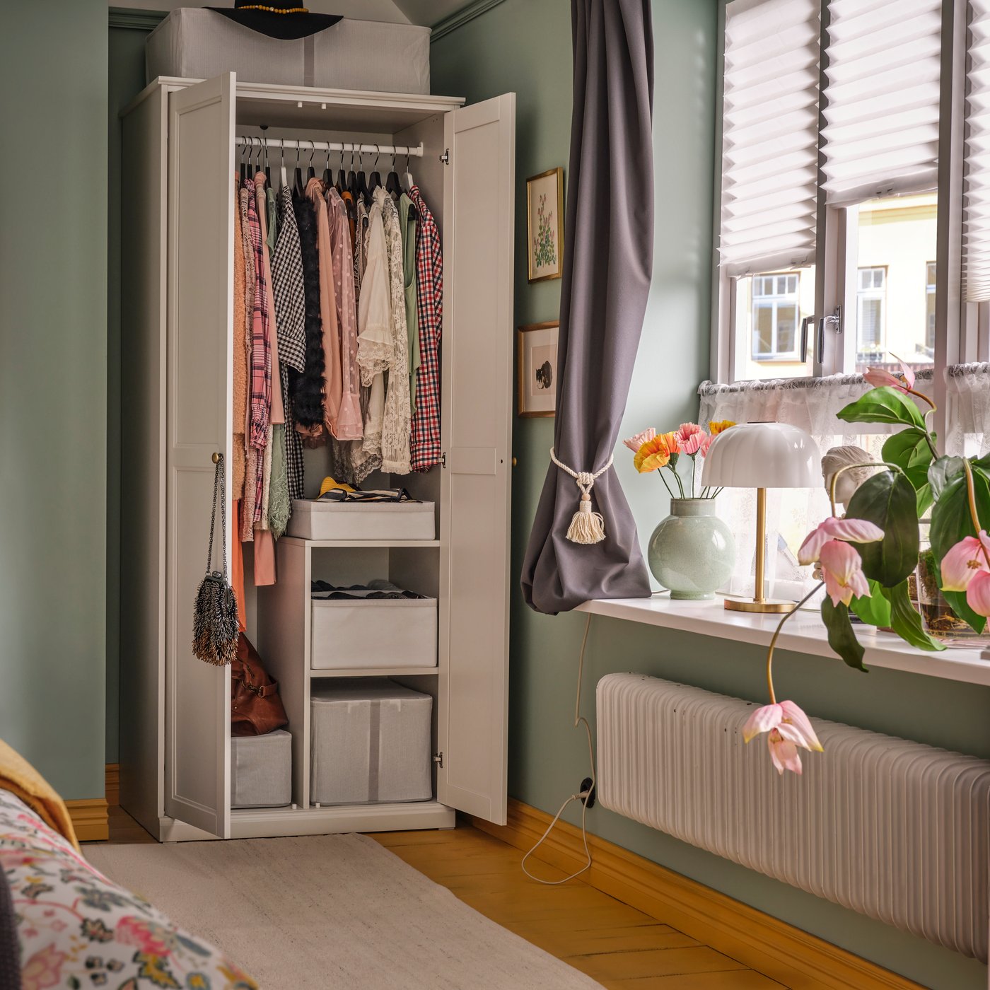 A bedroom with an off-white FÅGELFJÄLLET wardrobe containing clothes and storage boxes. It is next to a window with plants.