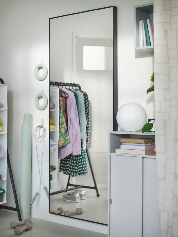 A large HOVET mirror with a black frame, reflecting a clothes rail with clothes. A grey cabinet, books and pastel gym gear.