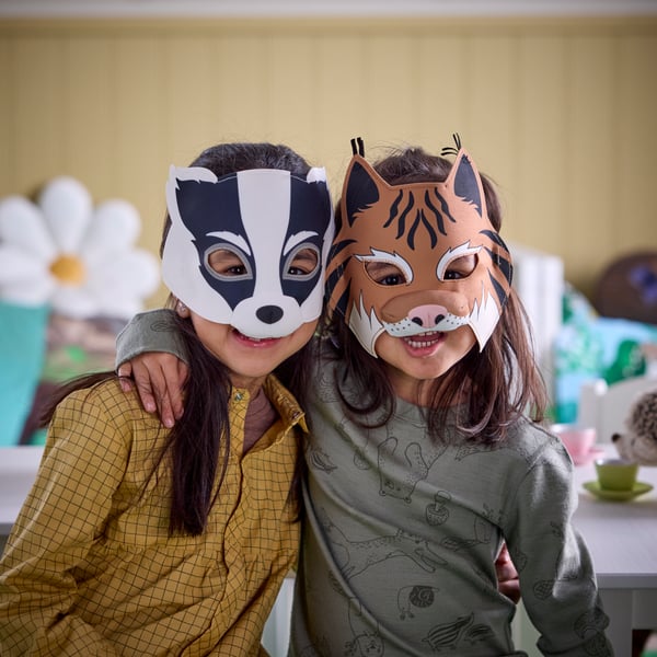 Zwei Kinder, die SKOGSDUVA Luchs- und Dachsmasken tragen, lächeln und halten sich gegenseitig um die Taille und die Schultern.