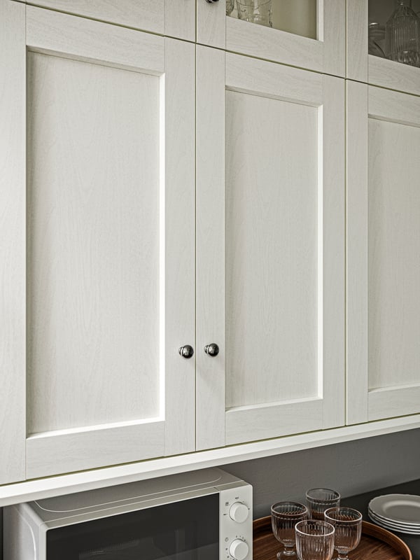 A SEKTION kitchen wall cabinet with ENKÖPING white kitchen front doors above a worktop with a microwave oven and glasses.