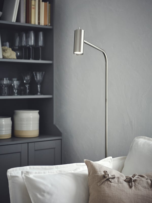 Dans un coin, un lampadaire VIRRMO nickelé allumé est posé entre un fauteuil BACKSÄLEN blanc et une armoire HAUGA grise.