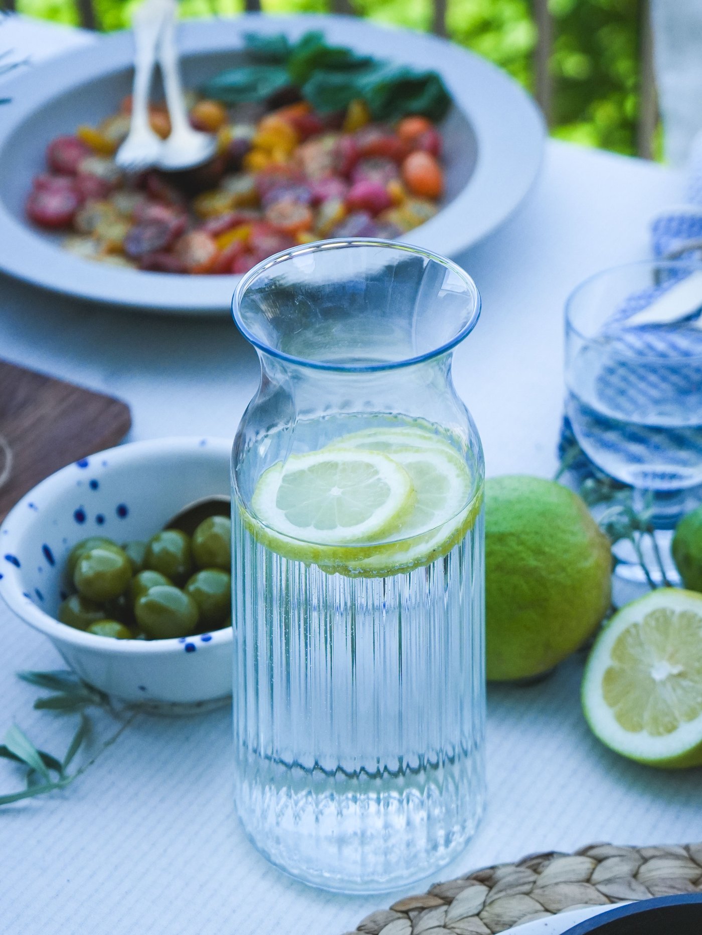 Tischdekoration mit einer VARDAGEN  Karaffe Zitronenwasser, grünen Oliven und einem Tomaten-Basilikum-Salat, umgeben von frischen Limetten und Zitronen.