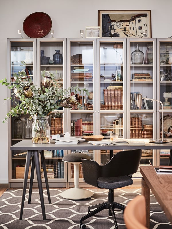 A NÄRSPEL desk with flowers in a vase and documents on top and three BILLY bookcases behind filled with books and mementos.