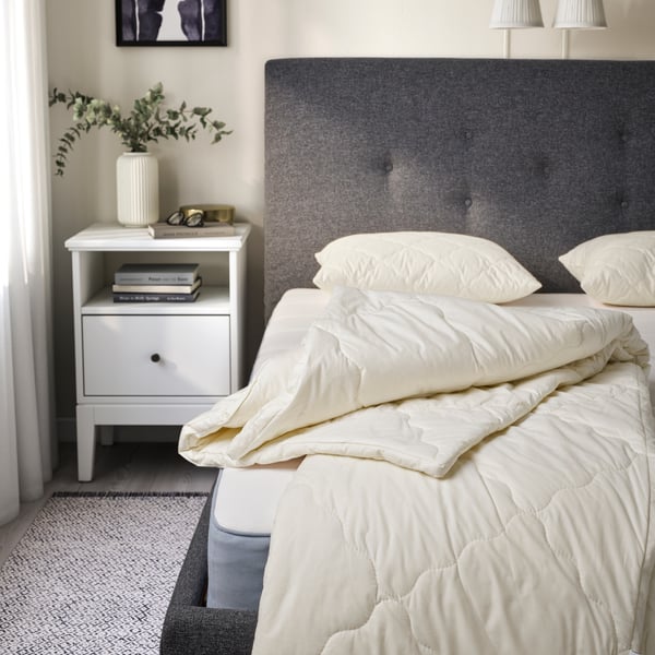 Eine weiße RÅDHUSVIN Bettdecke und weiße RÅDHUSVIN Kissen in einem dunkelgrauen Bett. Links vom Bett ein weißes Nachtkästchen.