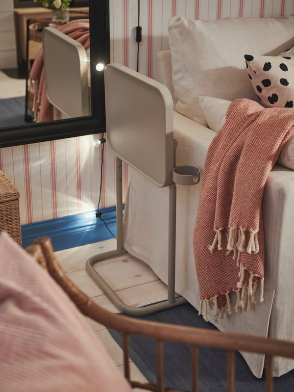 Table pour ordinateur portable BJÖRKÅSENA repliée à côté d’un canapé-lit HYLTARP avec un jeté ULLTISTEL posé sur le côté.