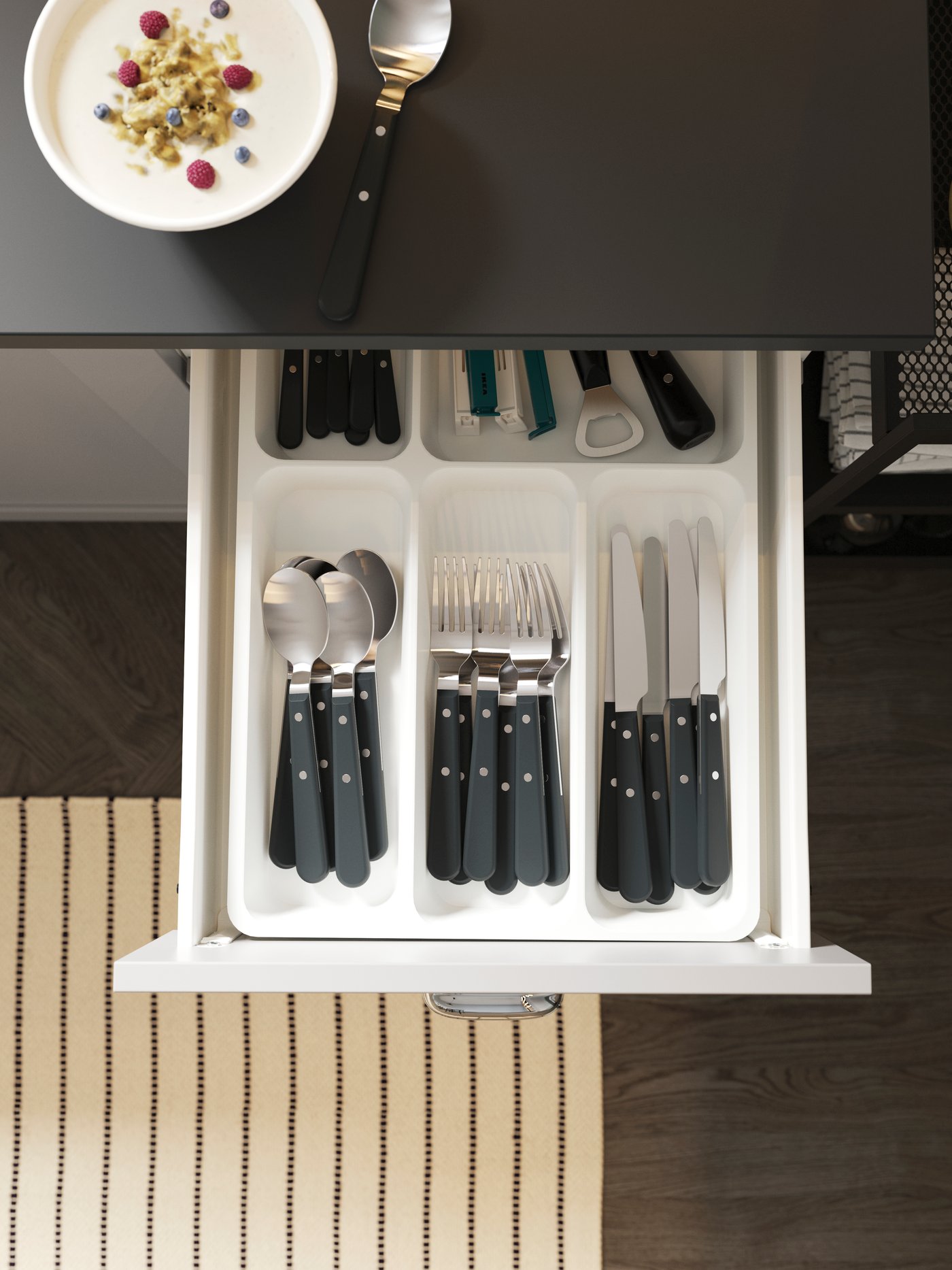 An open kitchen drawer with a cutlery tray neatly storing cutlery and kitchen utensils. A striped rug is on the floor below.