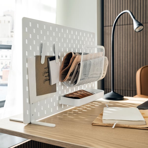 A wooden-surface desk with a white free-standing SKÅDIS pegboard on it that holds newspapers and accessories.