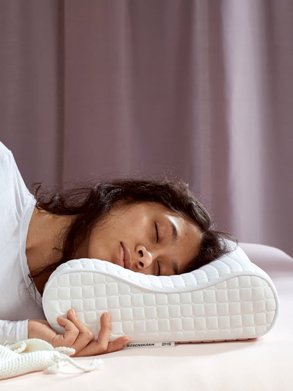 Una mujer recostada en una cama usa una almohada ergonómica que la ayuda a mantener su cuello descansado mientras duerme.