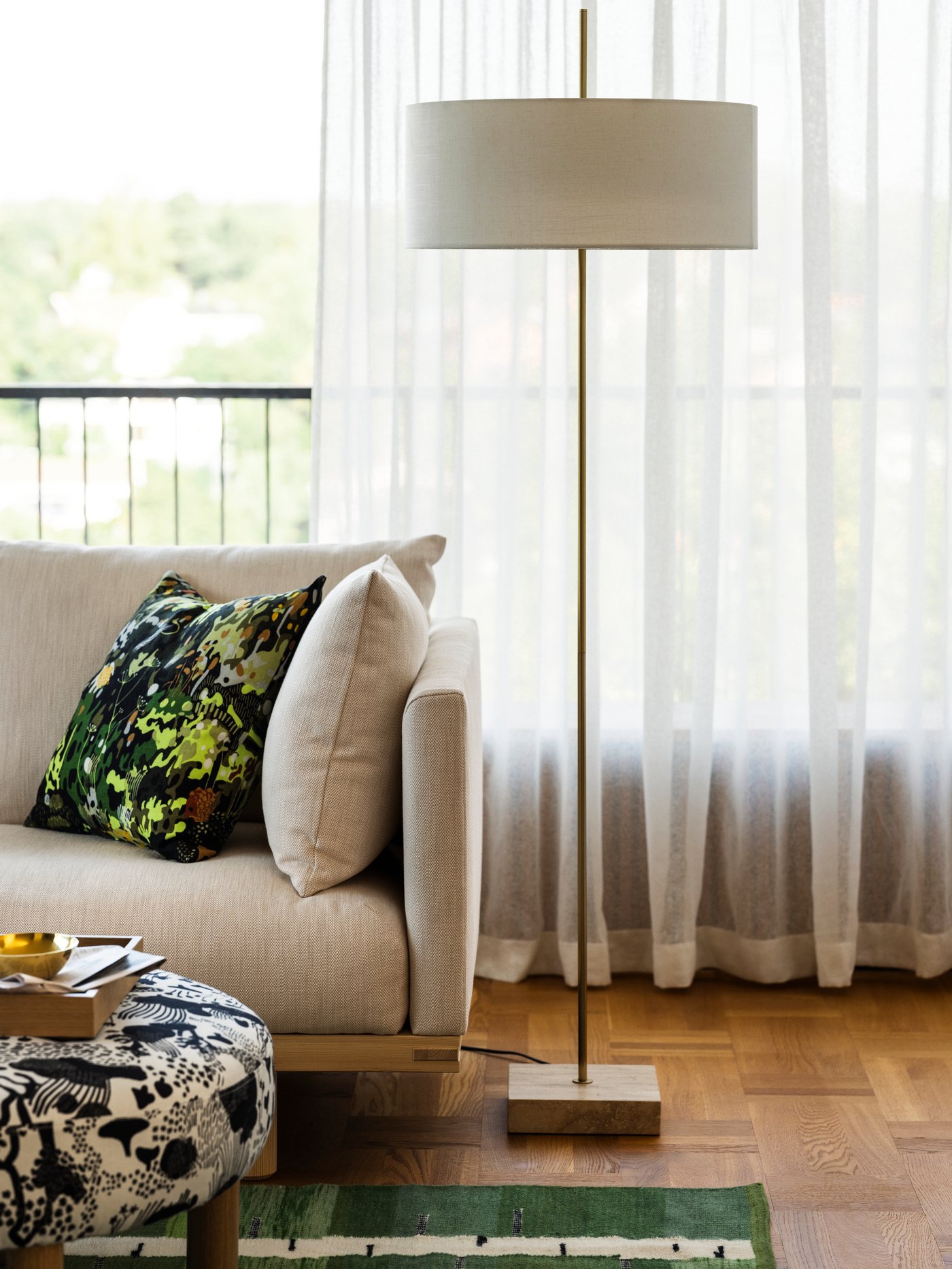 A brass-plated white STOCKHOLM 2025 floor lamp on parquet flooring, in front of white window curtains, with a sofa beside it.