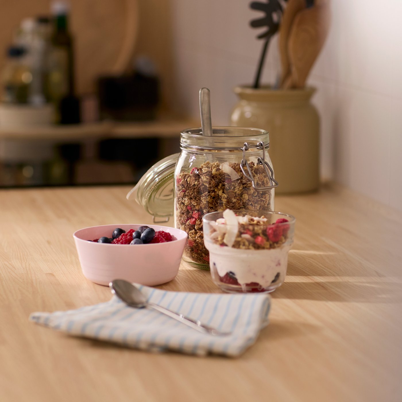 A glass jug with muesli in the back, and two smaller bowls filled with yoghurt and muesli ready to go.