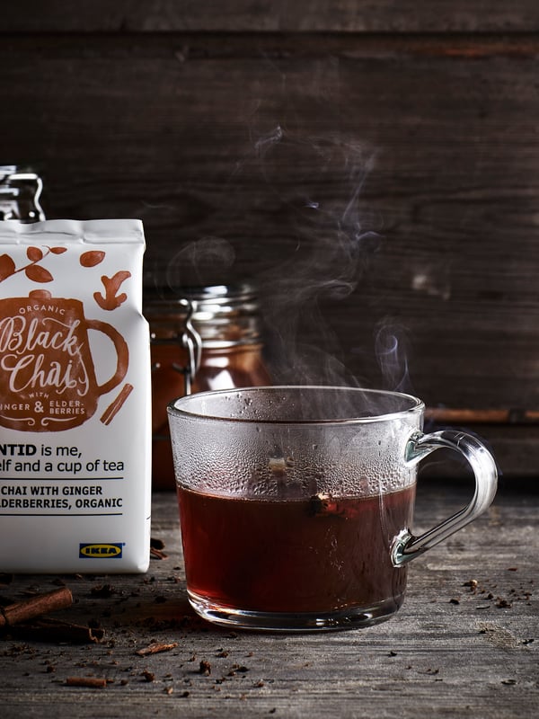 A cup of steaming EGENTID black chai tea with ginger and elderberries, on a wood table, by a packet of the tea.