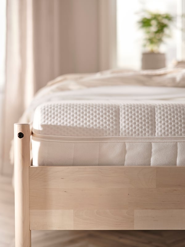 The end of a light wooden bed frame with an ÅKREHAMN foam mattress on it, in front of a window in a beige bedroom.
